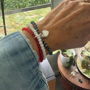 May include: A set of three beaded stretch bracelets worn on a wrist. The bracelets feature small, faceted beads in red, white, and dark gray. A small, polished silver-tone heart-shaped charm hangs from the dark gray beaded bracelet.