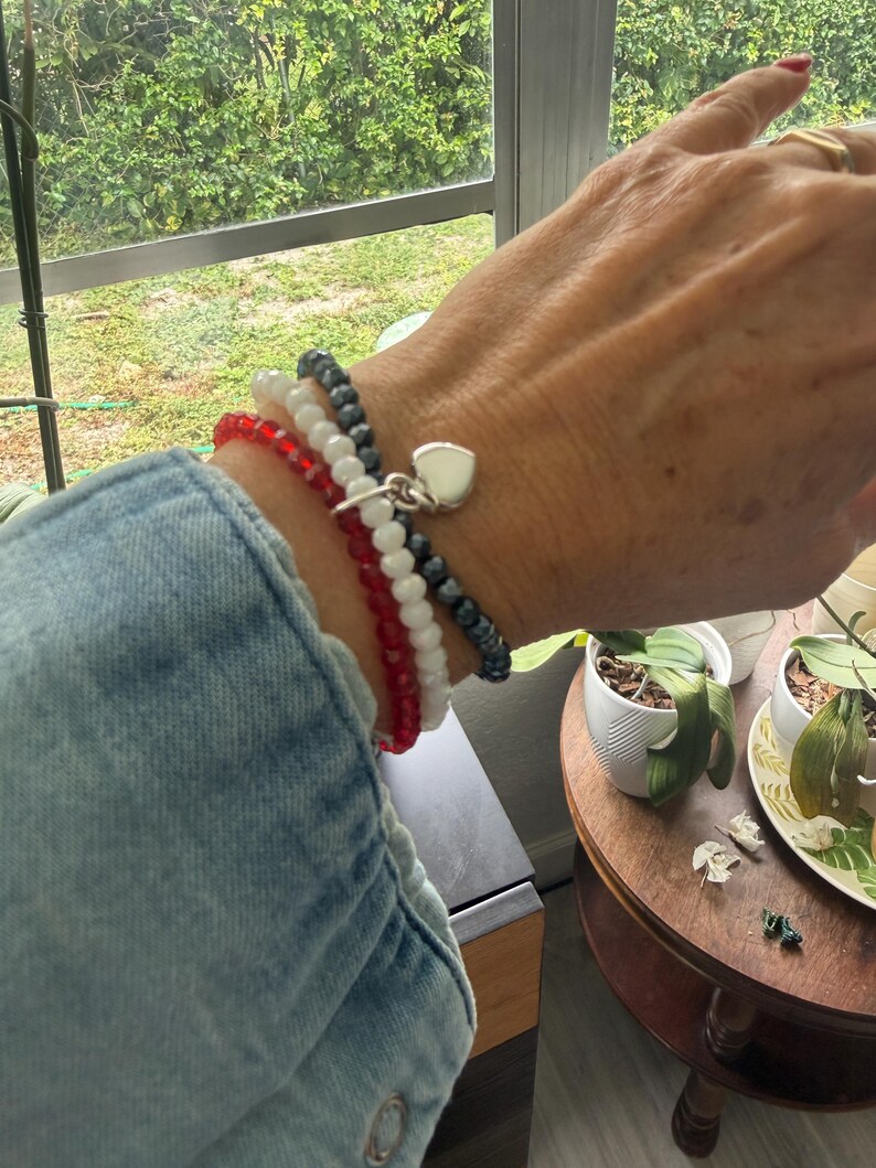 May include: A set of three beaded stretch bracelets worn on a wrist. The bracelets feature small, faceted beads in red, white, and dark gray. A small, polished silver-tone heart-shaped charm hangs from the dark gray beaded bracelet.