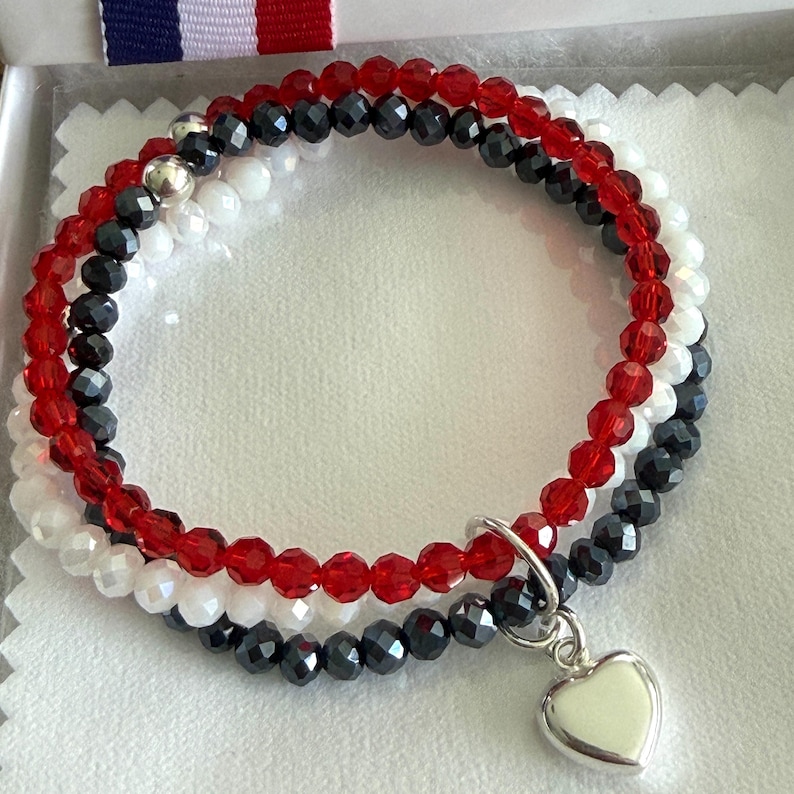 May include: A triple-strand beaded bracelet featuring faceted red, black, and white beads. The bracelet includes a silver-toned metal heart-shaped charm attached to the strands with a circular jump ring, creating a layered appearance.