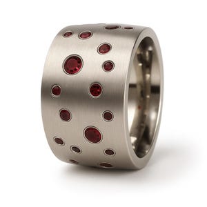 May include: A wide, brushed metal ring with numerous embedded red gemstones. The ring's surface is a light silver color, and the gemstones are various sizes and shapes. The ring is a bold piece of jewelry.