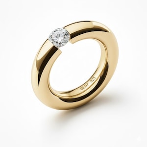 Könnte beinhalten: Ein goldener Ring mit einem breiten, abgerundeten Band und einem einzelnen, klaren Edelstein. Der Ring hat eine polierte Oberfläche und der Text "GEMP 925" ist auf der Innenseite des Bandes eingraviert. Weißer Hintergrund.