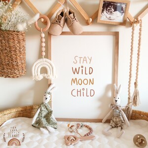 May include: A nursery decor print with the text "Stay Wild Moon Child" in a gradient of brown, orange, and yellow. The print is framed in a light wood frame and is hanging on a wall with a wooden rack above it. There are various baby items in the image, including a wooden bead garland, a crocheted rainbow, a pair of baby shoes, and two stuffed bunny rabbits.