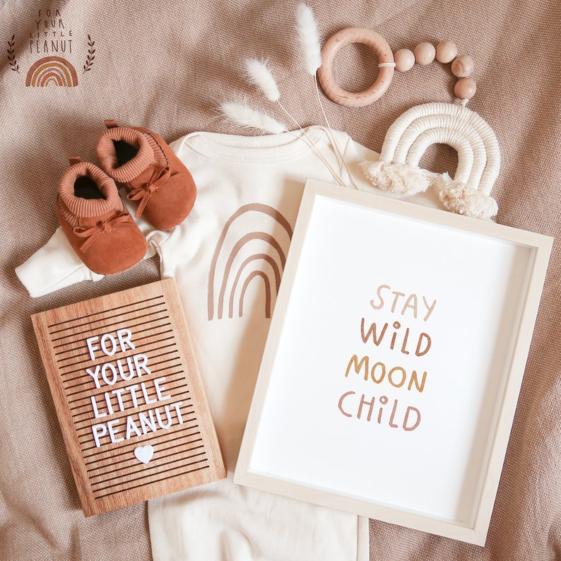 May include: A neutral-colored baby onesie with a brown rainbow design. A wooden letter board with the text "For your little peanut" and a white heart. A framed print with the text "Stay wild moon child" in brown, orange, and yellow. A wooden ring with brown beads and a white macrame rainbow hanging above.