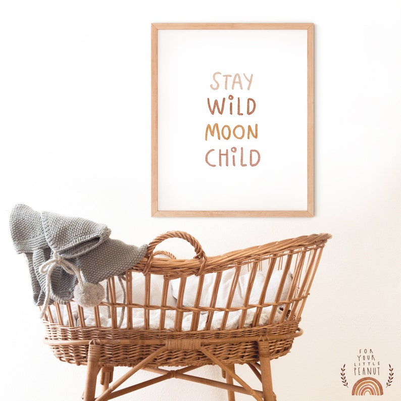 May include: A framed print with the text "Stay Wild Moon Child" in a light brown frame. The print is hanging on a white wall above a wicker bassinet.