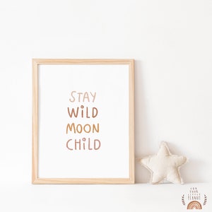 May include: A framed print with the text "STAY WILD MOON CHILD" in a light brown, orange, and pink color scheme. The print is in a wooden frame and is sitting on a white surface.
