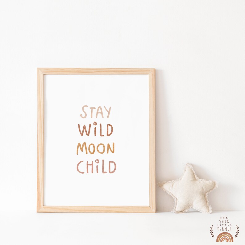 May include: A framed print with the text "STAY WILD MOON CHILD" in a light brown, orange, and pink color scheme. The print is in a wooden frame and is sitting on a white surface.