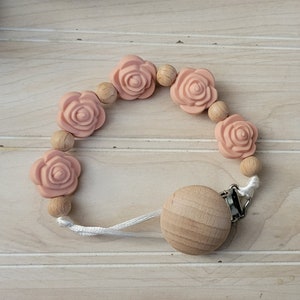 May include: A pink silicone and wooden baby teether with a rose design. The teether has a wooden clip and a white string.