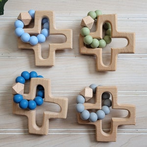 May include: Four wooden cross-shaped teethers with silicone bead rings in different colors: light blue, green, blue, and gray.