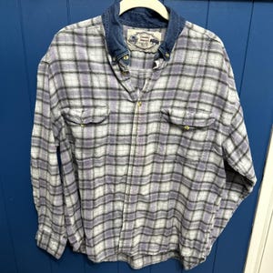 May include: A long-sleeved, button-down shirt with a plaid pattern in shades of lavender, white, and black. The shirt features a denim collar and two chest pockets. The label on the collar reads "Double Impact".