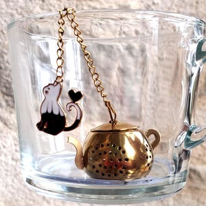 Könnte beinhalten: Ein goldfarbener Tee-Ei in Teekannenform mit perforiertem Körper, befestigt an einer goldenen Kette mit einem schwarz-weißen Katzenanhänger und einem kleinen Herzdetail, platziert in einer durchsichtigen Glastasse mit Henkel.
