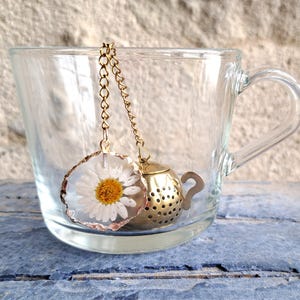 Puede incluir: Una taza de té de vidrio transparente contiene un infusor de té dorado con forma de tetera pequeña y una margarita encapsulada en resina. El infusor cuelga de una delicada cadena. La taza de té está sobre una superficie de madera azul.