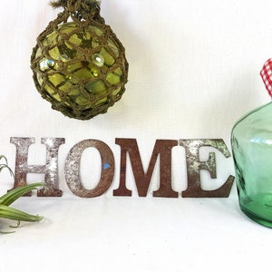 Op de afbeelding: Het woord "HOME" is gespeld in grote, roestige metalen letters. Een groene glazen fles met een rood-wit geruite hartdecoratie bevindt zich aan de rechterkant van de afbeelding. Een groene glazen drijver hangt bovenaan de afbeelding.