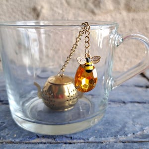 Può includere: Una tazza da tè in vetro trasparente con un infusore per tè color oro. L'infusore ha un piccolo cestino a forma di teiera e un ciondolo decorativo a forma di ape con accenti gialli e neri. Il ciondolo ape è sospeso all'interno della tazza.