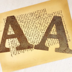 Op de afbeelding: Twee grote, roestige metalen letters, "A" en "A", liggen op een vel oud, vergeeld papier met handgeschreven tekst. De letters zijn licht verweerd en hebben een rustieke uitstraling.