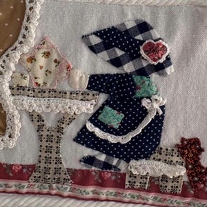 May include: A fabric wall hanging featuring a patchwork scene with a figure in a navy blue polka dot dress and a checkered hat. The scene includes a floral teapot on a table, a brown curtain with white dots, and a red floral border at the bottom.