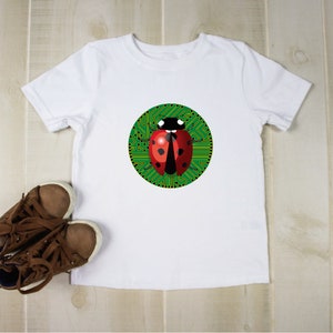 Può includere: T-shirt bianco con un disegno di coccinella rossa e nera su sfondo di circuito stampato verde. Scarpe da ginnastica marroni sono posizionate vicino alla maglietta su una superficie di legno chiaro. La maglietta è probabilmente per bambini.