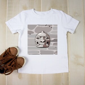May include: White t-shirt featuring a graphic of a stylized mask with a stone wall background. Brown shoes with white soles are next to the shirt. The mask has a light gray and brown color scheme.
