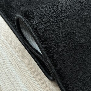 May include: Close-up of a black rug with a textured surface and a black border. The rug is folded over, revealing a glimpse of the lighter-colored backing. The rug is on a light wood-grain surface.