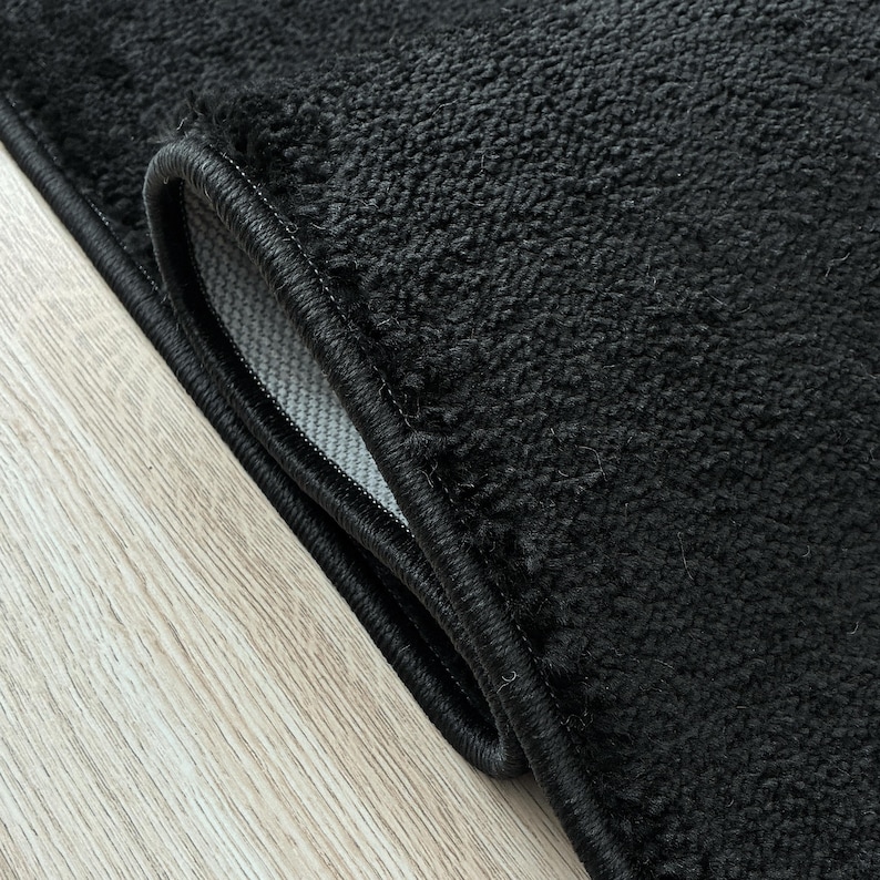 May include: Close-up of a black rug with a textured surface and a black border. The rug is folded over, revealing a glimpse of the lighter-colored backing. The rug is on a light wood-grain surface.