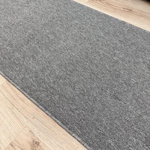 May include: A gray runner rug with a textured surface. The rug is rectangular and has a narrow, dark gray border. The rug is placed on a light-colored wooden floor.
