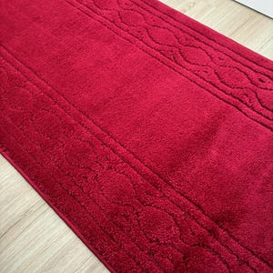 May include: A rectangular deep red bath mat featuring a plush, textured surface. The mat has a solid center panel bordered by two distinct bands of raised, repeating oval patterns that run along the length of the rug on both sides.