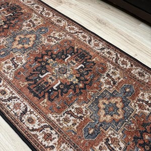 May include: A rectangular runner rug with an ornate, traditional design. The rug features a central pattern in shades of rust, navy, and cream, with a black border. The rug is placed on a light-colored wooden floor.