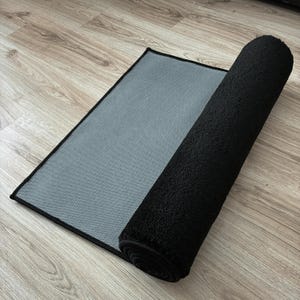 May include: A black rectangular rug with a rolled-up section. The rug has a textured, plush surface and a gray, woven backing. The edges are neatly finished with a black border. The rug is partially rolled, revealing the backing.