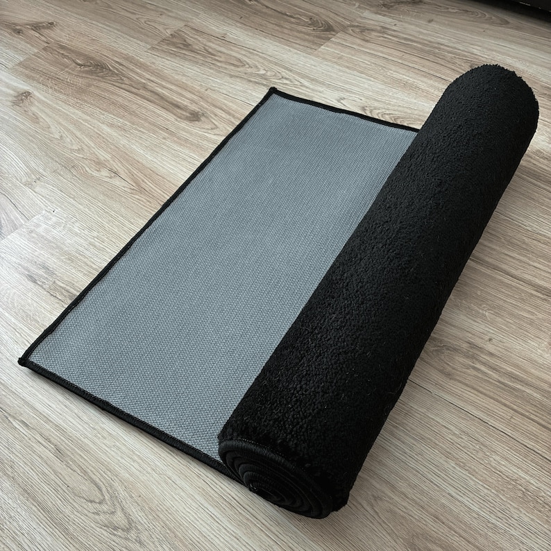 May include: A black rectangular rug with a rolled-up section. The rug has a textured, plush surface and a gray, woven backing. The edges are neatly finished with a black border. The rug is partially rolled, revealing the backing.