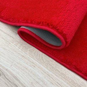 May include: Close-up of a rolled-up red rug with a gray backing. The rug has a plush texture and a red border. The rug is on a light wood surface.