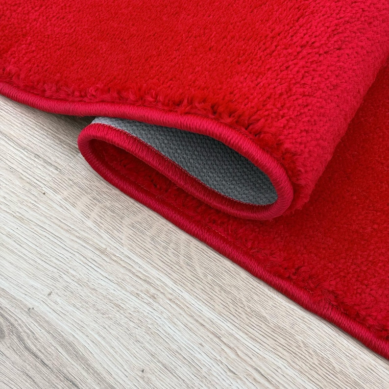 May include: Close-up of a rolled-up red rug with a gray backing. The rug has a plush texture and a red border. The rug is on a light wood surface.