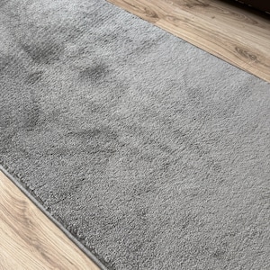 May include: A rectangular, gray rug with a textured surface. The rug is placed on a wooden floor. The rug's edges are neatly finished with a thin border.
