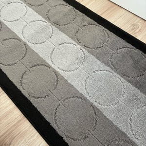 May include: A rectangular runner rug featuring a repeating pattern of interconnected circles. The design consists of a central light gray stripe flanked by medium gray sections, all bordered by solid black edges on both long sides.