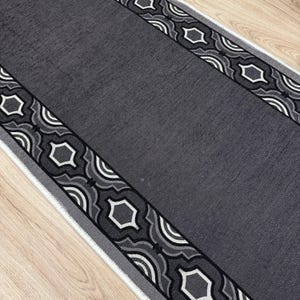 May include: A gray runner rug with a geometric border design. The border features repeating patterns of white, black, and gray shapes. The rug is rectangular and appears to be made of a soft material.