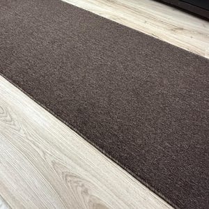 May include: A rectangular runner rug featuring a solid dark brown color with a short, dense, and uniform pile texture. The edges of the rug are finished with a matching dark brown stitched binding that runs along the entire length of the piece.