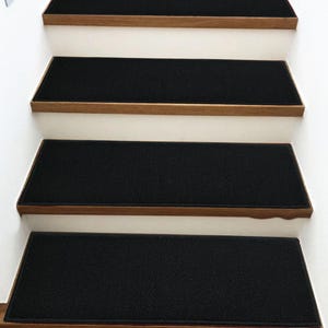 May include: Four rectangular black stair treads with a textured surface are installed on wooden stair steps. Each tread covers the horizontal surface of the step, contrasting with the white vertical risers of the staircase.