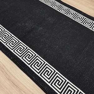 May include: A black runner rug with a white Greek key pattern border. The rug has a textured surface and is placed on a light-colored wooden floor. The design is symmetrical, with the pattern running along both edges of the rug.