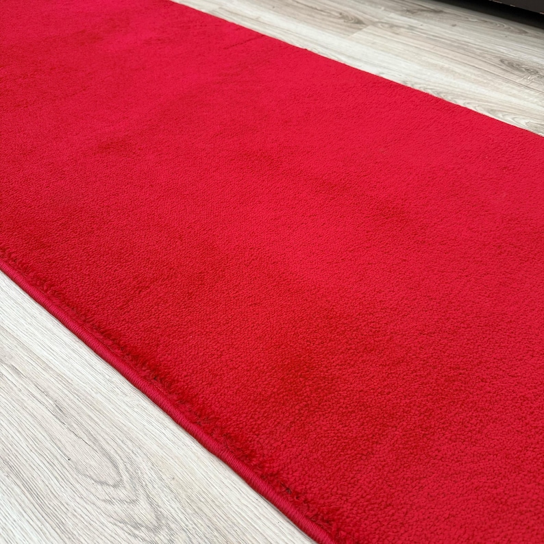 May include: A vibrant red rectangular rug with a soft, textured surface. The rug is a solid color and appears to be made of a plush material. The edges are neatly finished, and the rug is placed on a light-colored wooden floor.