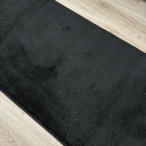 May include: A rectangular black rug with a soft, plush texture. The rug is a solid black color and appears to be made of a dense material. The rug is placed on a light-colored wooden floor.