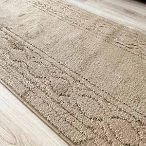May include: A rectangular beige area rug featuring a textured, raised pile surface with a repeating geometric border pattern. The rug has a soft, tufted appearance with a consistent light tan color throughout the entire fabric construction.