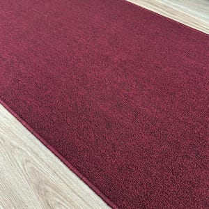 May include: A rectangular burgundy rug with a textured surface. The rug has a dark red border and is placed on a light wood floor. The rug is a solid color and appears to be made of a soft material.
