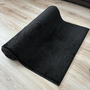 May include: A rolled-up, rectangular black rug with a textured surface. The rug is made of a soft material and has a dark border. The rug is partially unrolled, revealing its plush texture.