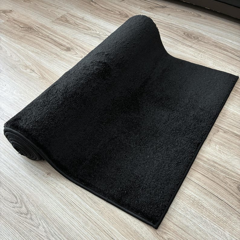 May include: A rolled-up, rectangular black rug with a textured surface. The rug is made of a soft material and has a dark border. The rug is partially unrolled, revealing its plush texture.