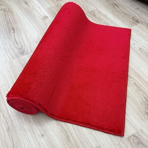May include: A rolled-up, bright red rectangular rug with a textured surface. The rug is partially unrolled, revealing its plush texture and a contrasting wooden floor background. The rug's edges are neatly finished.