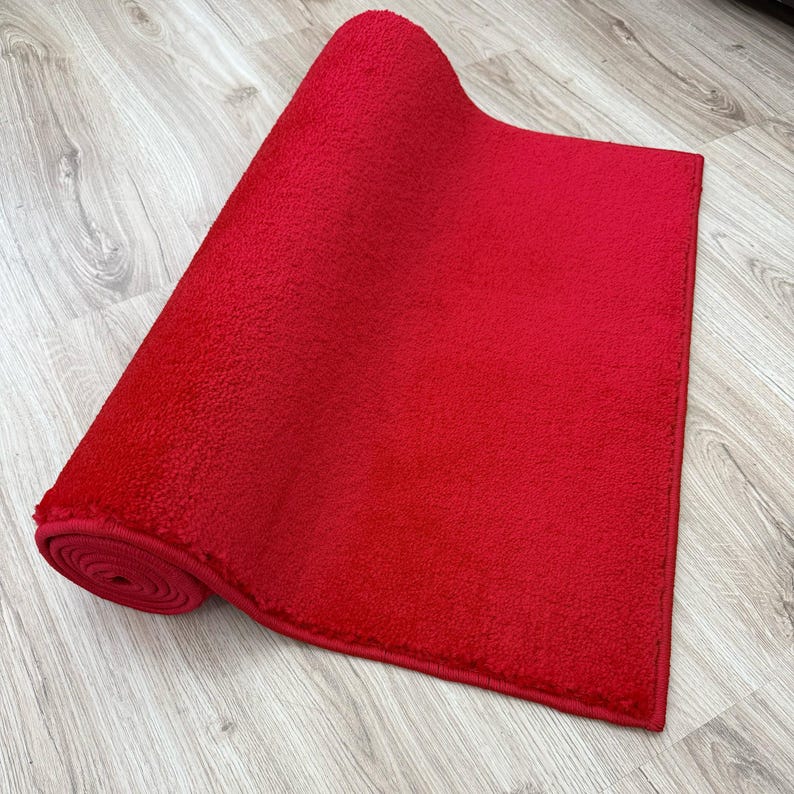 May include: A rolled-up, bright red rectangular rug with a textured surface. The rug is partially unrolled, revealing its plush texture and a contrasting wooden floor background. The rug's edges are neatly finished.