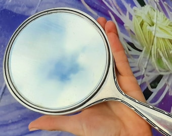 May include: A silver hand mirror with a round, bevelled glass surface and a decorative handle. The mirror is held in a hand against a purple and white floral background.