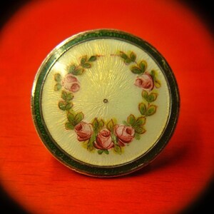 May include: A round, white enamel pin with a green border. The pin features a floral design with pink roses and green leaves.