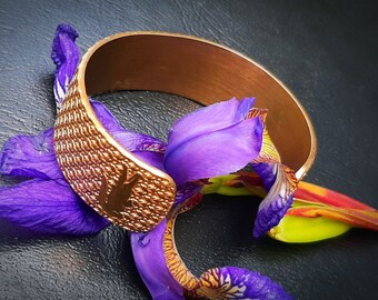 May include: A copper bracelet with a textured surface and a silhouette of a bird design. The bracelet is resting on a purple and yellow flower.