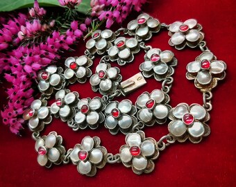May include: A silver-toned necklace featuring flower-shaped links with mother-of-pearl petals and red center stones. The necklace is displayed on a red velvet surface, with pink flowers in the background.