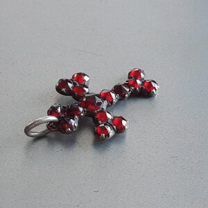 May include: A silver cross pendant with red garnet stones. The cross is made of metal and has a loop at the top for attaching to a chain.