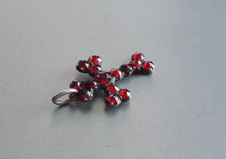 May include: A silver cross pendant with red garnet stones. The cross is made of metal and has a loop at the top for attaching to a chain.